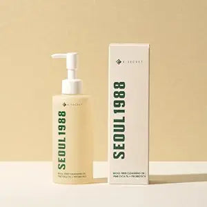 A bottle of k-secret 1988 cleansing oil with a pump dispenser next to its product box on a light beige surface. The bottle and the box feature the brand name “K-Secret” and the product name “Seoul 1988” in a distinctive vertical olive-green font.
